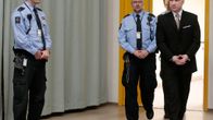 Breivik saapuu saliin puku päällä, taustalla vartijoita.