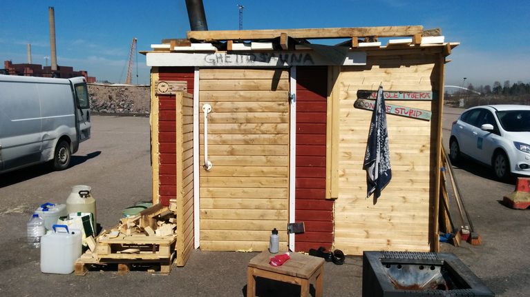 Hökkelin näköinen, lautarakenteinen väliaikainen sauna. Rakennuksen edessä vesiastioita ja polttopuita. Seinässä lukee "ghettosauna" ja "Älä ole typerä".