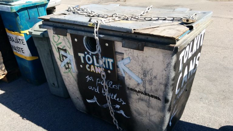 Harmaa roskalaatikko, jonka ympäri on sidottu kettingit. Laatikon kyljessä lukee tölkit.
