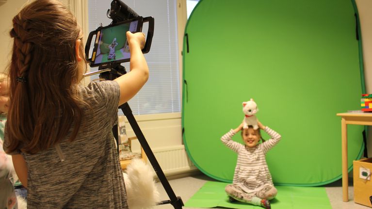 Päiväkotilapset kuvaavat toisiaan tabletilla green screen-kankaan edessä.