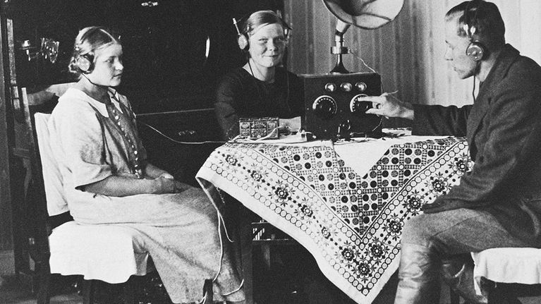 Radionkuuntelua 1920-luvulla.Kaksi naista ja mies pöydän ääressä kuuntelemassa radiota (kuulokkeet).  