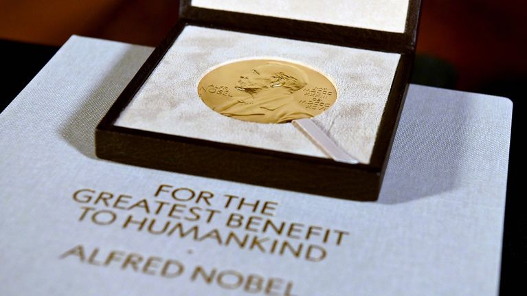 Nobel-mitali laatikossa. Alla on levy, jossa lukee englanniksi "ihmiskunnan suurimmaksi hyödyksi, Alfred Nobel"