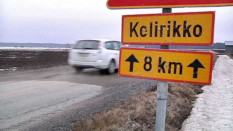 Valkoinen henkilöauto ajaa kelirikon kourissa olevaa soratietä. Ympärillä olevilla pelloilla on laikuttain lunta.