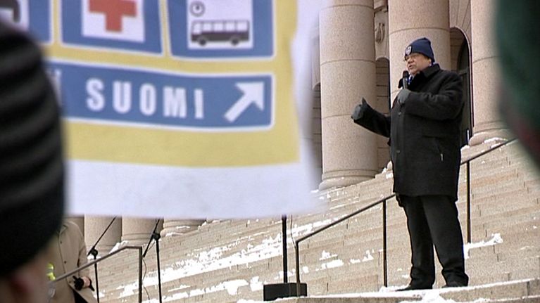 Timo Soini pitää puhetta eduskuntatalon portailla helmikuun lopussa 2011, etualalla liehuu perussuomalaisten vaalibanderolli.