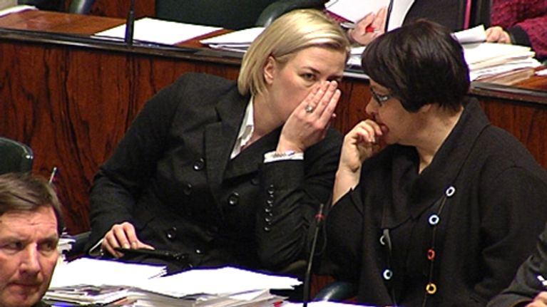 Jutta Urpilainen kuiskaa vieressään istuvalle Tarja Filatoville eduskuntasalissa käsi suunsa edessä.