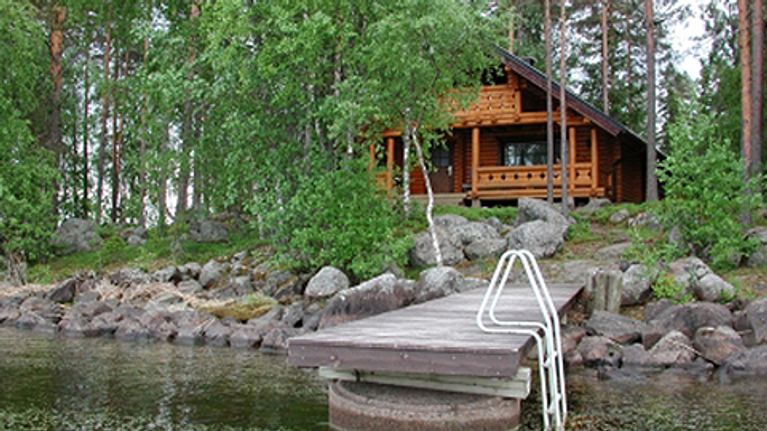 Kuvassa hirsinen huvila kivikkoisella järvenrannalla kesäisessä maisemassa, etualalla laituri.