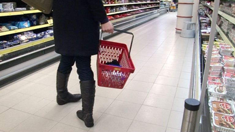 Nainen kantaa ostoskoria kaupan käytävällä.