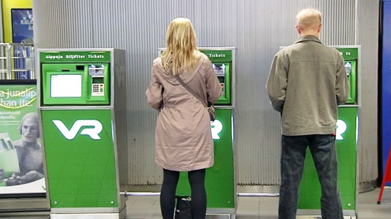 Mies ja nainen ostavat junalippuja automaateista rautatieasemalla.