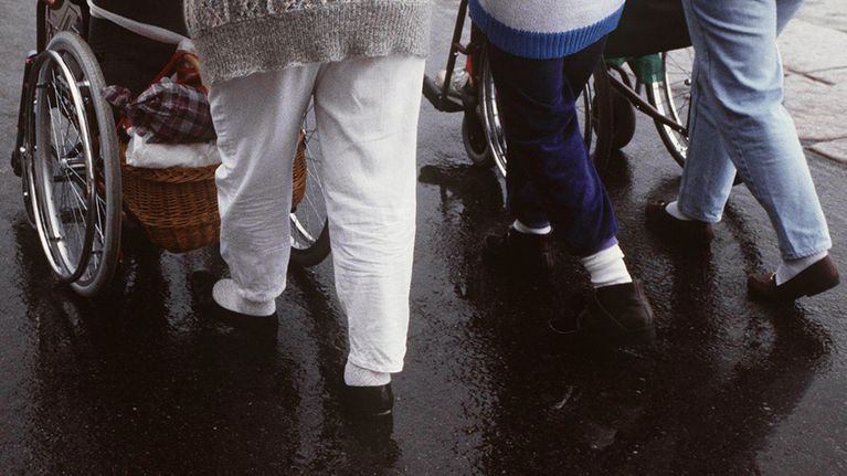 Hoitajat työntävät liikuntavammaisia pyörätuoleissa.