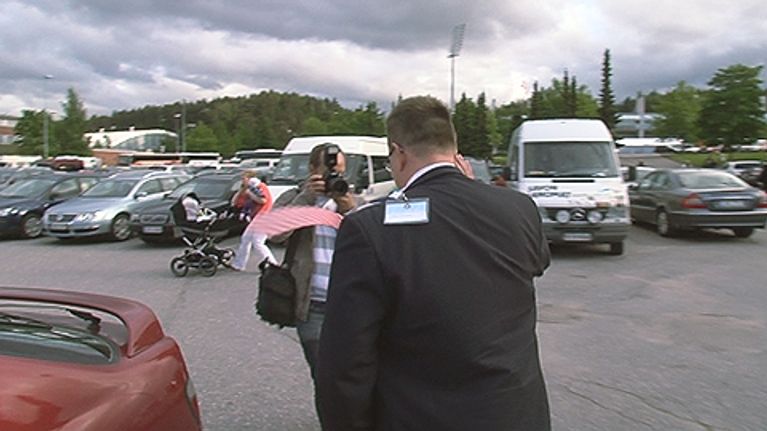 Mies kävelee parkkipaikalla ja toinen ottaa tästä kuvaa