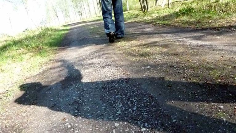 Kuvassa näkyy hiekkaista polkua kulkevan jalat, takana vaanii tuntemattomaksi jäävän henkilön varjo.