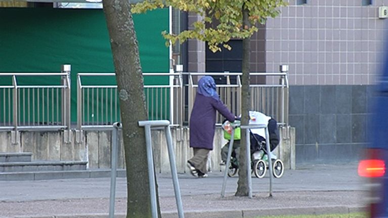 Maahanmuuttajanainen työntää lastenvaunuja kadulla. Auto ajaa ohi.