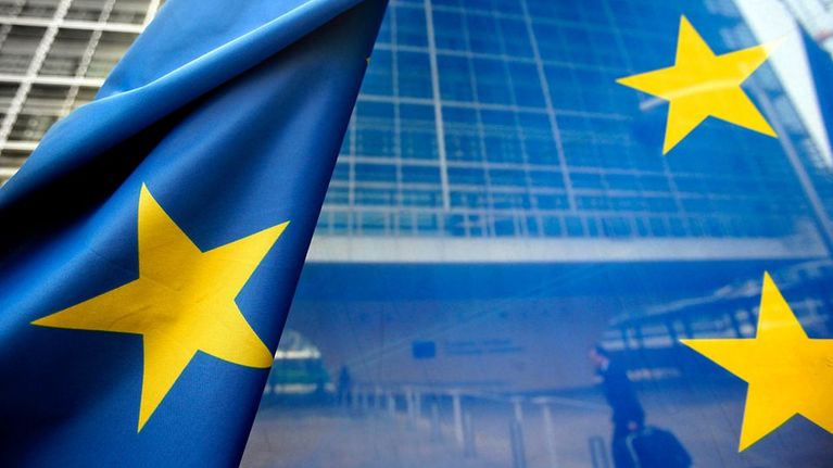 EU:n lippu heiluu Euroopan Unionin rakennusten edessä Brysselissä. Lipun läpi näkyy matkalaukkua vetävä mies.