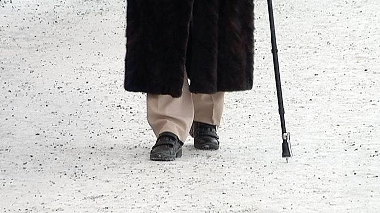 Vanhuksen jalat ja kävelykeppi hiekoitetulla jalkakäytävällä.