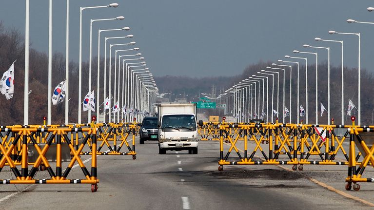 Kuvassa kaksi kulkuneuvoa ajaa keltamustien porttien välistä, ajotien vierellä liehuu Etelä-Korean lippuja.