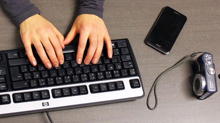 Naisen kädet tietokoneen näppäimistöllä. Pöydällä älypuhelin ja digikamera.