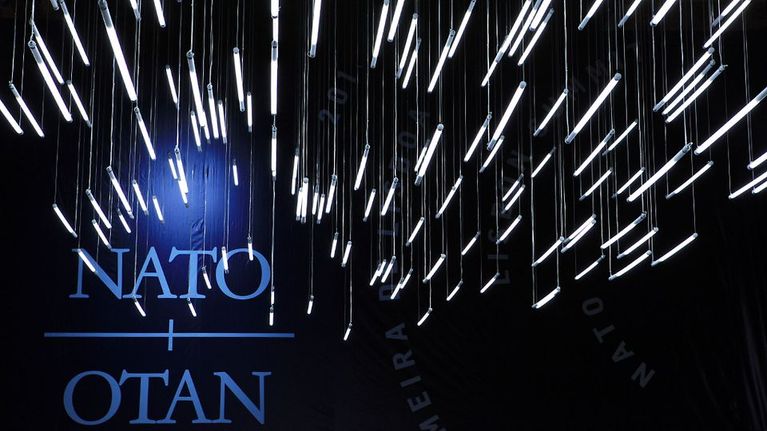 Katosta riippuu lukuisia valkoisia loistelamppuja mustaa taustaa vasten. Takaseinällä NATO / OTAN -tekstit.