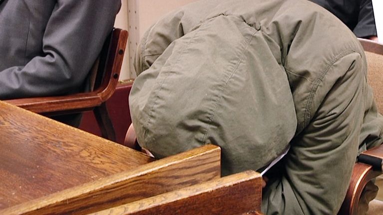 Mies on vetänyt vihreän maiharin hupun kasvojensa suojaksi ja nojaa päätään oikeussalin pöytään.
