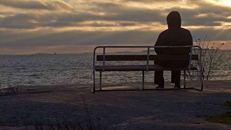 Henkilö istuu puiston penkillä pimenevässä illassa ja tuijottaa merelle. 