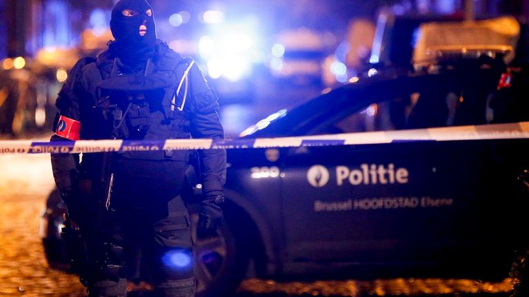 Raskaissa suojavarusteissa oleva poliisi, jolla on päässään musta hiihtomaski, seisoo poliisin sinivalkoisen eristysnauhan takana. Miehen takana näkyy poliisiauto, jonka kyljessä lukee 'politie'.