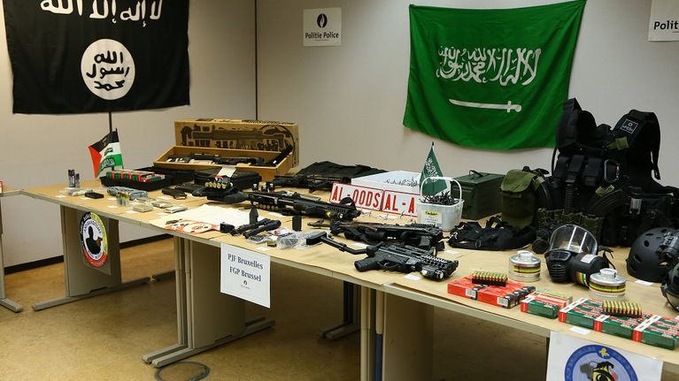 Pöydällä on lukuisia erilaisia aseita ja ammuksia sekä suojavarusteita. Seinälle on ripustettu islamistien lippuja. Vasemmalla on musta lippu, jossa on valkoista arabiankielistä kirjoitusta ja mustaa kirjoitusta valkoisessa ympyrässä, ja oikealla vihreä lippu, jossa on valkoista arabiankielistä kirjoitusta.