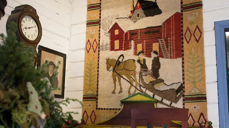 Seinällä olevan ryijy, jossa kuva kahdesta henkilöstä hevosreen kanssa lumisessa maisemassa. 