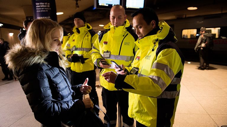 Kirkkaankeltaiseen vartijan asuun pukeutunut mies tarkistaa vaalean sinitakkisen naisen henkilöllisyystodistusta. Taustalla näkyy kaksi muuta samanlaisiin keltaisiin pusakoihin pukeutunutta miestä.