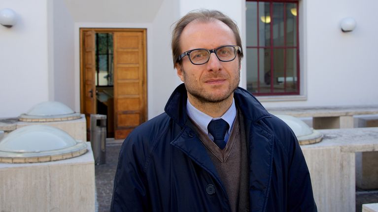 Silmälasipäinen dosentti Gino Scaccia sinisessä trenssitakissaan valkoisen rakennuksen luona.
