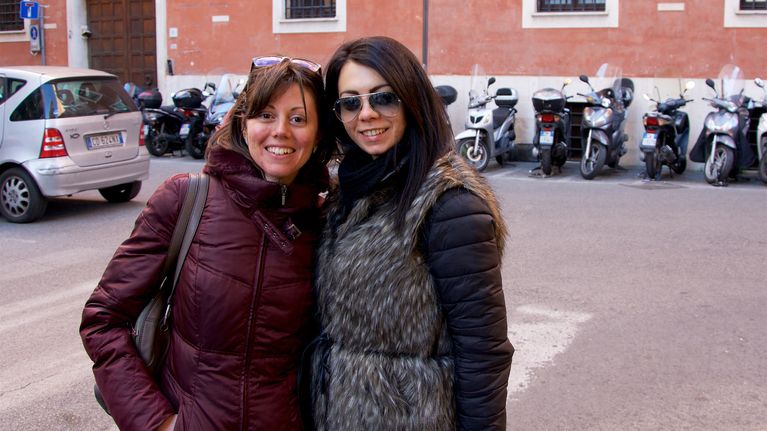 Hymyilevät roomattaret rakennuksen edessä, jonka edustalle on parkkeerattu skoottereita. Elisalla aurinkolasit otsalla ja viininpunainen toppatakki. Pamelalla lentäjämalliset aurinkolasit päässä. Hän on pukeutunut harmaaseen tekoturkisliiviin ja mustaan toppatakkiin.