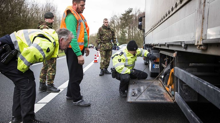 Kolme huomioliiveihin pukeutunutta poliisia tutkii rekka-autoa maantiellä. Kaksi maastopukuista sotilasta seuraa vieressä. 