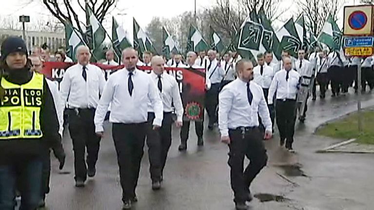 Valkopaitaisten ja mustakravattisten miesten rivistö kantaa vihreä-valkoisia lippuja. Edessä kulkee poliisi. 