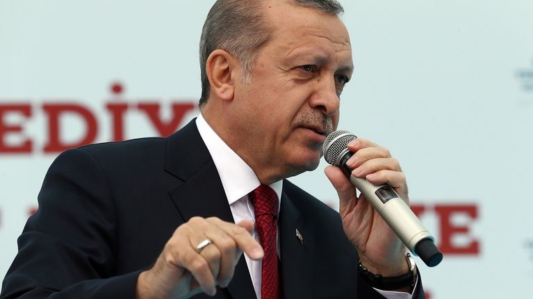 Erdoğan puhuu vasemmassa kädessään olevaan mikrofoniin ja osoittaa oikean kätensä etusormella alaspäin. Hänellä on tumma puku, punainen ruudullinen kravatti. 