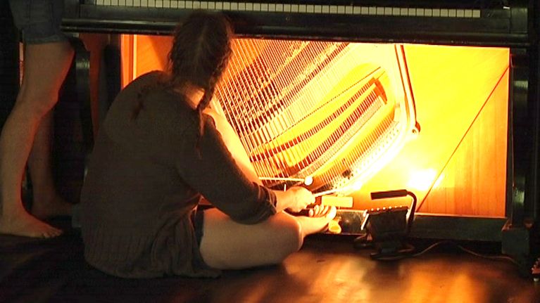 Nainen istuu pianon edessä, jonka sisukset on paljastettu. Hän soittaa pianoa pianon sisältä.