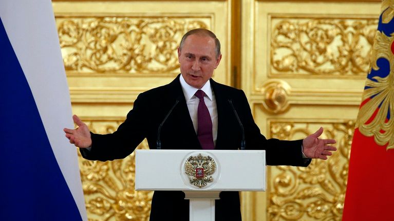 Putin puhuu korokkeen takaa ja levittää juuri käsiään. Putinilla on tumma asu ja viininpunainen kravatti. Taustalla näkyy suuri kullattu ja koristeltu ovi ja kuvan oikeassa ja vasemmassa reunassa Venäjän liput. Korokkeessa on Venäjän vaakunaeläin, kaksipäinen kotka.