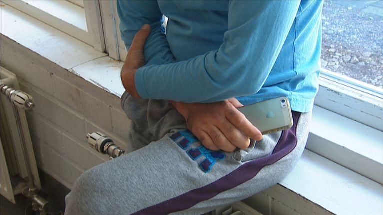 Anonyymi turvapaikanhakija nojaa ikkunalautaan kännykkä kädessään.