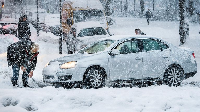 Mies lapioi lunta auton edestä.