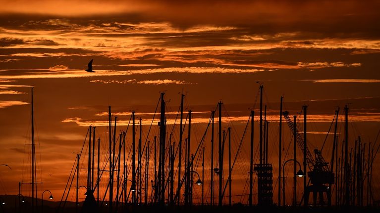 Oranssinpunainen auringonnousu veneiden mastojen takana. 