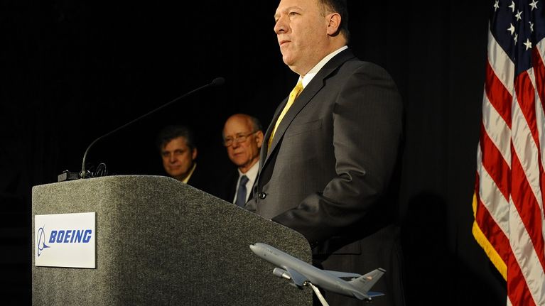 Mike Pompeo seisoo puhujankorokkeen ääressä tummassa puvussa, keltaisessa kravatissa ja katsoo vakavana yleisöön. Taustalla on Yhdysvaltain lippu ja muita henkilöitä.