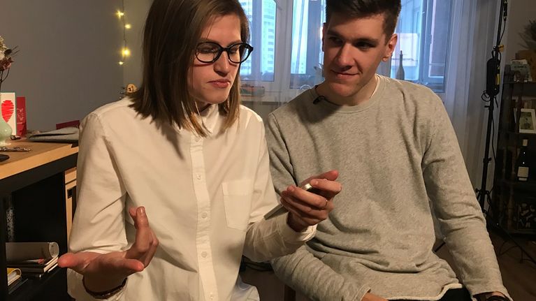 Nuori nainen valkoisessa kauluspaidassa, vieressä istuu nuori mies harmaassa trikoopaidassa.