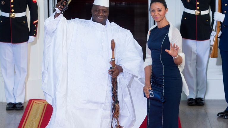 Jammeh ja hänen puolisonsa seisovat punaisella matolla ja tervehtivät ihmisiä hymyillen.