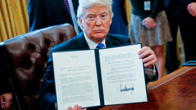 Kirjoituspöydän ääressä istuva Trump pitää käsissään ohutta kansiota, jossa on allekirjoitettu paperi.