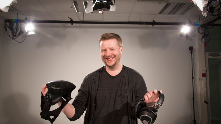 Mies poseeraa studiossa virtuaalilasit ja kuulokkeet kädessä.