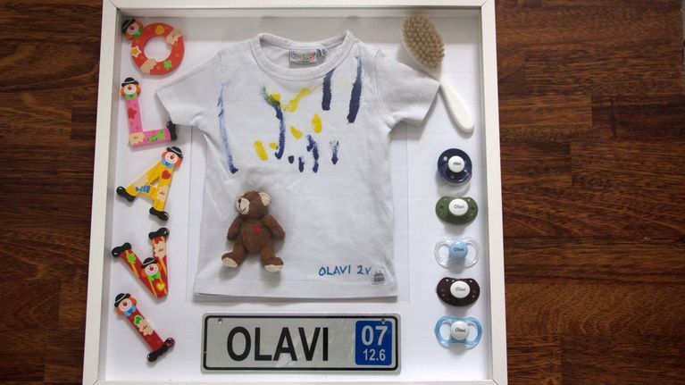 Muistotaulu, jossa lapsen t-paita, tutteja ja hiuskarja.