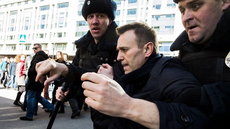 Kaksi poliisimiestä karvalakeissa kuljettaa Navalnyia kainaloista nostaen. Taustalla näkyy ihmisjoukkoa ja auringon kirkkaasti valaisema kerrostalon seinä. Navalnyi on paljan päin, mustassa takissa.