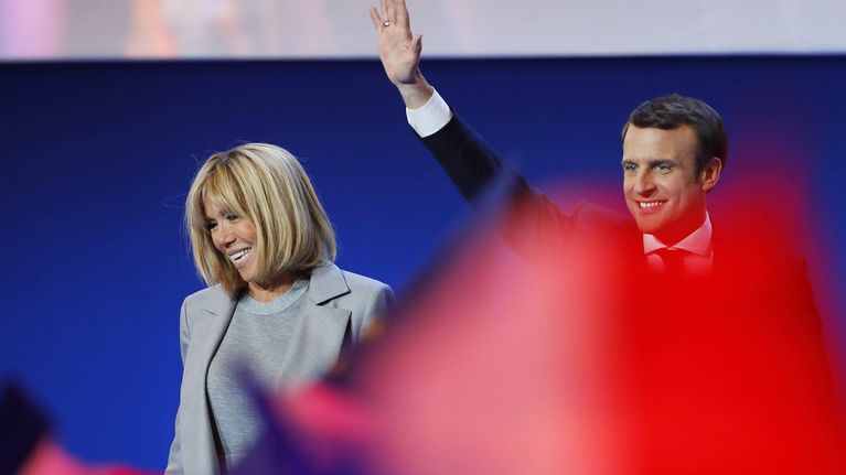 Brigitte Trogneux ja Emmanuel Macron seisovat esiintymislavalla. Macron heiluttaa kättään. Etualalla joku heiluttaa jonkinlaista punaista vaatetta, joka näkyy kuvassa punaisen värin leiskahduksena.