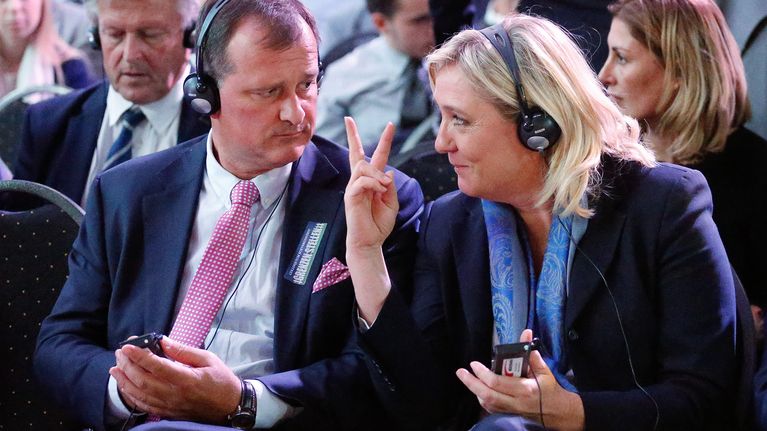 Louis Alliot ja Marine Le Pen istuvat kokoontumisessa. Molemmilla on päässään kuulokkeet. Le Pen näyttää juuri Aliot'lle oikean kätensä etu- ja keskisormea.