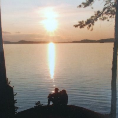 Kaksi ihmishahmoa istuvat kalliolla veden äärellä ihailemassa auringonlaskua.