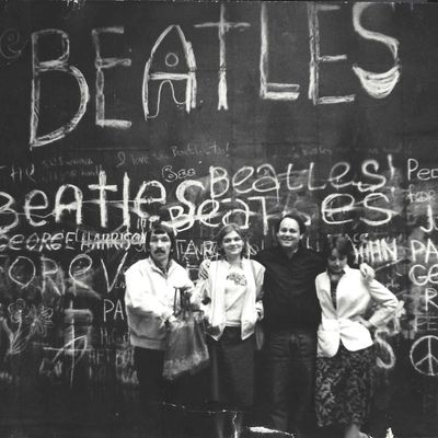 Neljä nuorta aikuista poseeraa iloisesti seinän edessä. Seinään on kirjoitettu The Beatles -yhtyeen jäsenten nimiä ja piirretty rauhanmerkkejä.